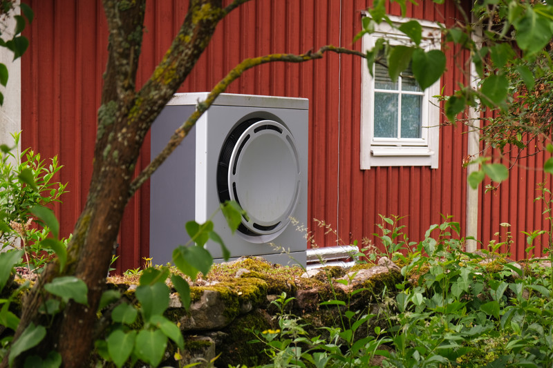 Grå värmepump installerad utanför ett rött trähus med vita fönster, omgiven av grön sommarvegetation.